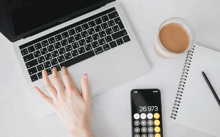 A person typing on a laptop with a smartphone calculator app open beside a cup of coffee