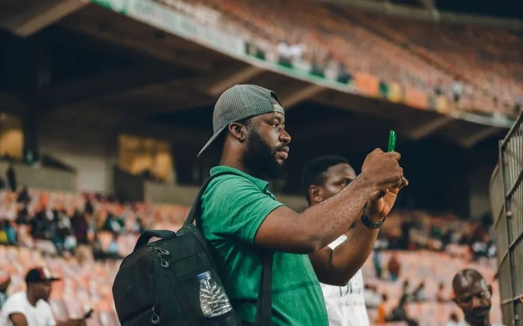 Man in a green shirt taking a photo with his smartphone from packed stadium seating during a night game