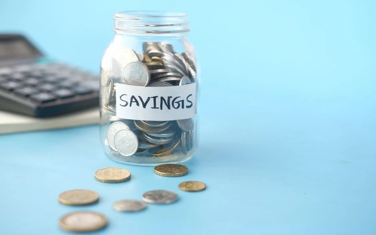 Glass jar labeled Savings filled with coins next to a calculator on a light blue desk