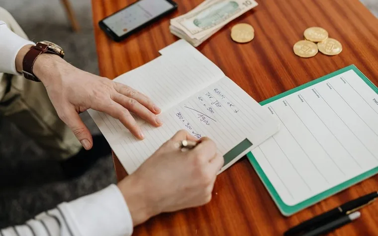 Hands writing calculations in a notebook next to cash on a wooden desk