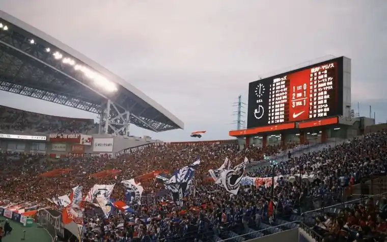 Crowded stadium at night with fans waving flags beneath an illuminated scoreboard