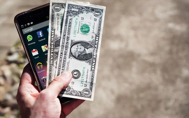 Hand holding US dollar bills next to a smartphone
