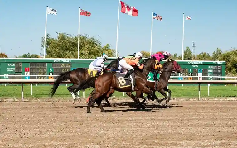 Jockeys in colorful silks racing horses down a dirt track with flags and a green tote board in the background