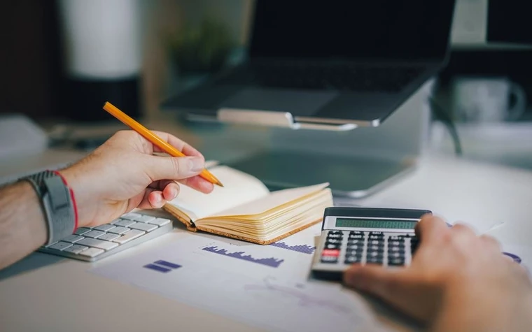 Hand holding a pencil over an open notebook next to a calculator and laptop on a desk