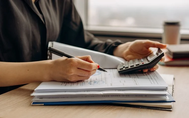 Hand holding a calculator over a stack of paperwork on a desk, with a pen and coffee cup nearby