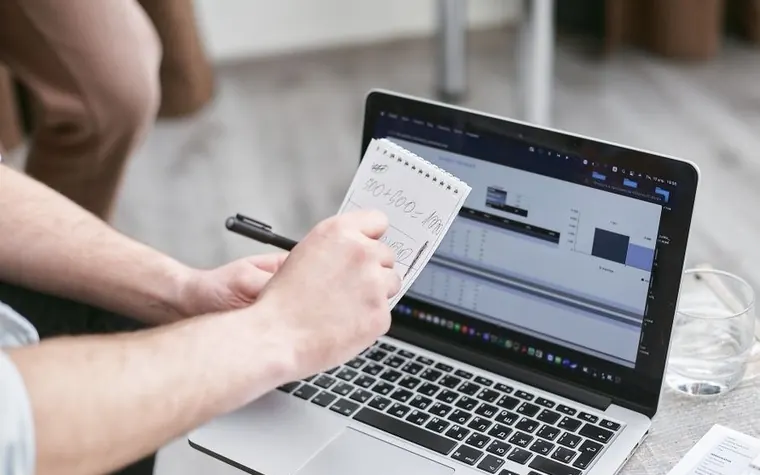 A person writing an addition sum on a notepad while a laptop shows a spreadsheet with charts