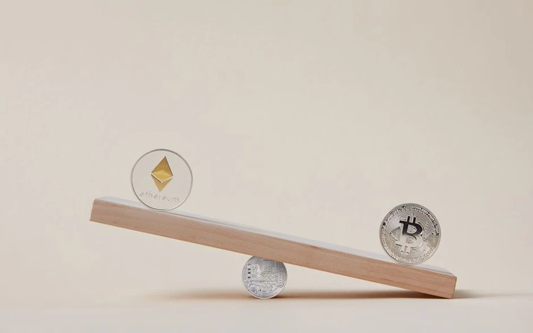 Two silver coins stamped with Ethereum and Bitcoin symbols balanced on opposite ends of a small wooden seesaw