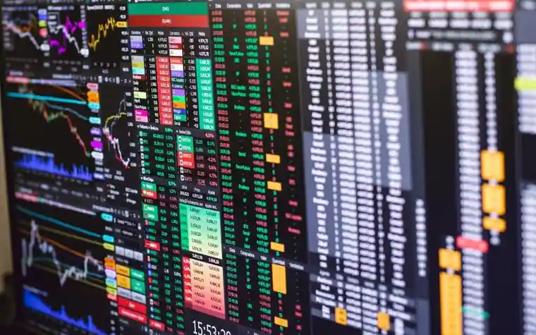 A wall of financial trading screens displaying colorful stock price charts and scrolling data tables