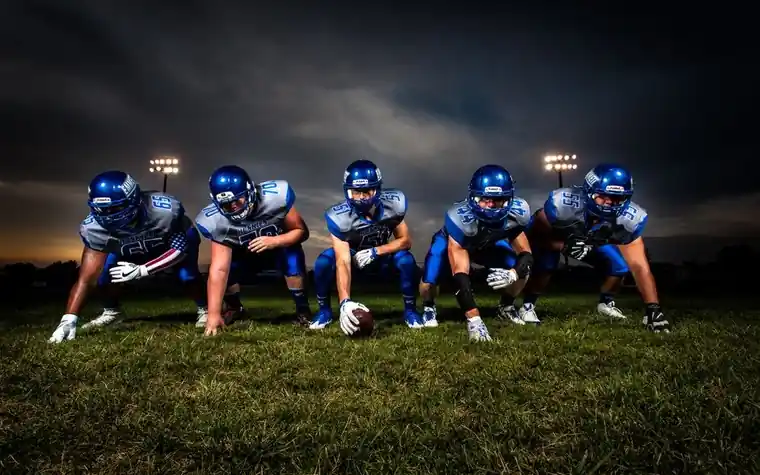 Five football players in blue uniforms crouched in an offensive line formation under stadium lights at night