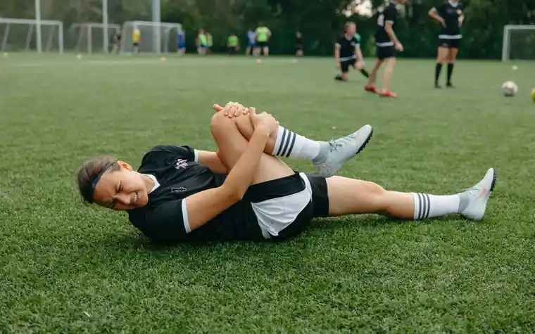 A young soccer player lying on the grass holding her knee in pain during a match while play continues in the background