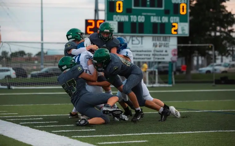 High school football players in green helmets making a group tackle in front of a scoreboard reading zero to zero