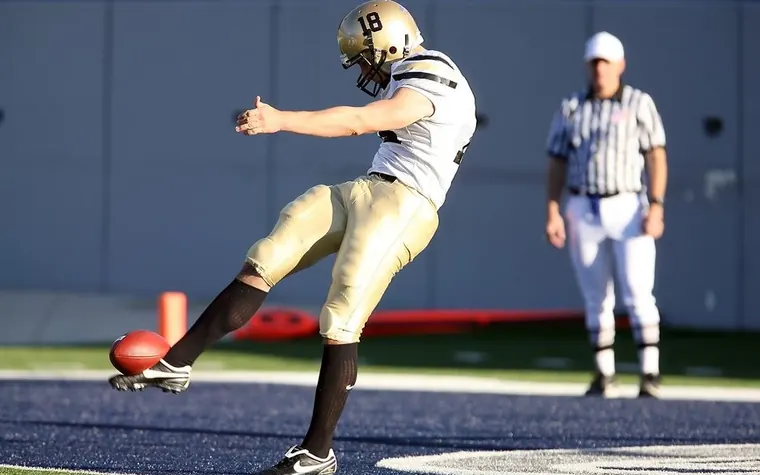 A football kicker in a gold and white uniform punting the ball downfield as a referee watches
