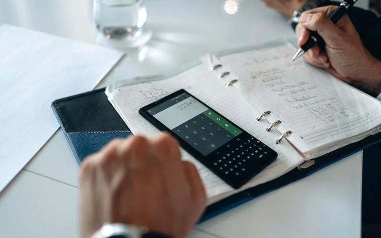 A hand holding a smartphone calculator app open next to a paper planner covered in handwritten figures
