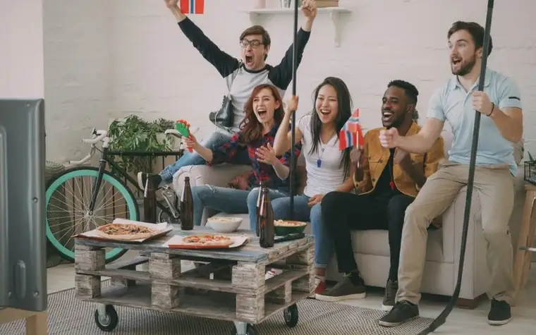 Group of friends on a sofa cheering with their arms raised while watching a game on television