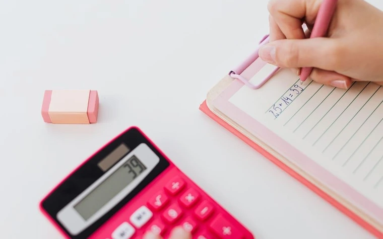 Hand writing figures in a lined notepad next to a pink calculator on a white desk