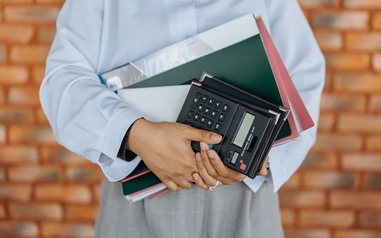 A person in an office shirt holding a calculator and folders against a brick wall background