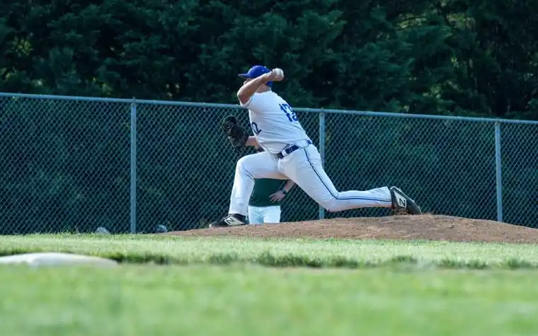 Baseball pitcher mid-throw on the mound during a game with a chain-link outfield fence behind him