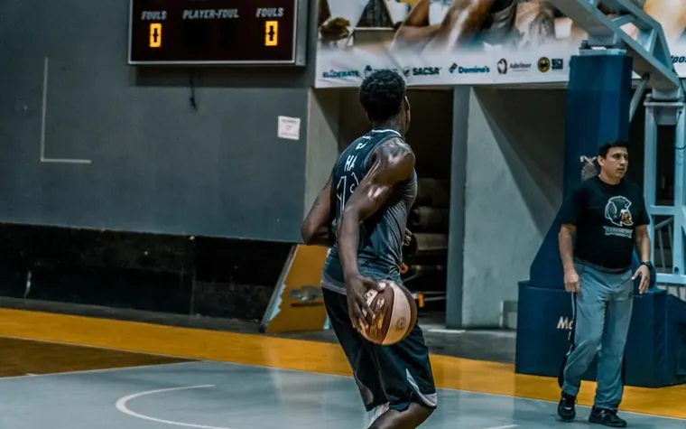Basketball player dribbling downcourt in an arena with a lit scoreboard showing fouls and period count on the wall
