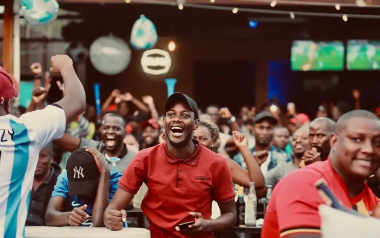 Crowd of fans cheering in an outdoor bar with multiple TV screens showing different live games in the background