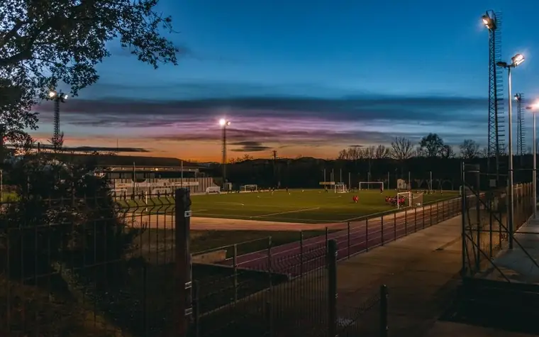 Small soccer stadium under floodlights at dusk with a match in progress and a purple sunset sky