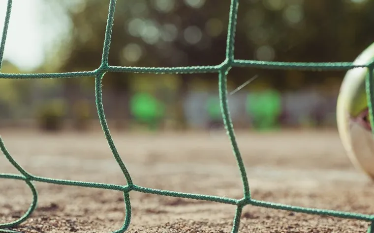 Ground-level view of a green goal net with a soccer ball wedged against it on a dirt pitch