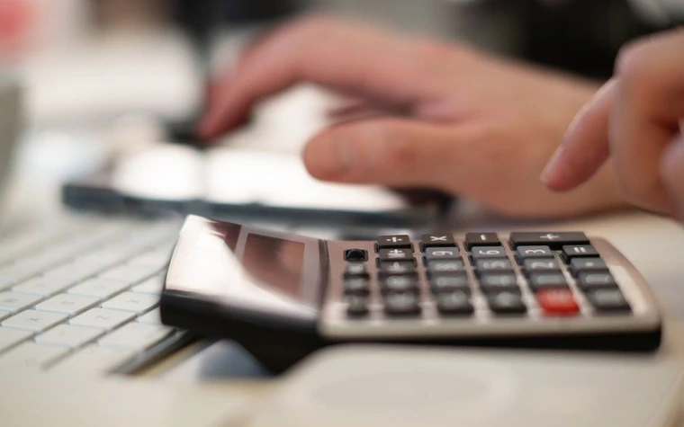 Close-up of a hand pressing buttons on a calculator next to a keyboard and phone on a desk