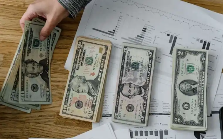 Overhead view of a hand sorting US dollar bills next to printed financial charts on a desk
