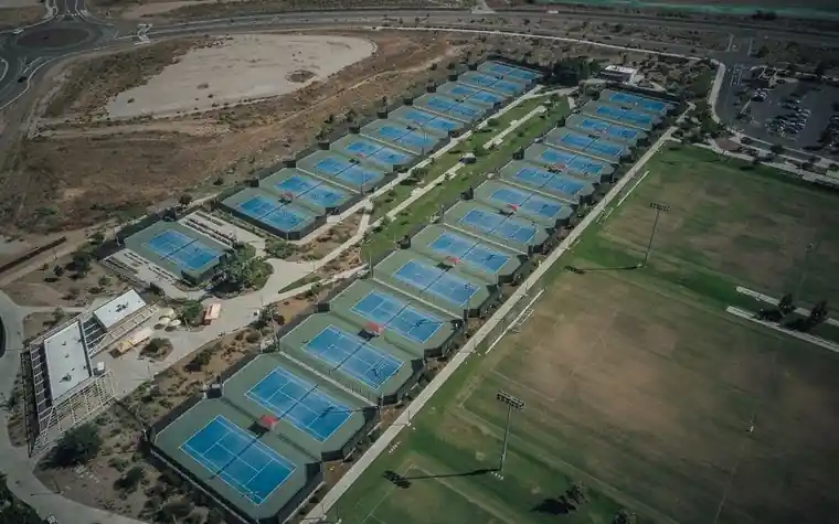 Aerial view of a large tennis complex with dozens of blue and green courts arranged in rows next to open playing fields