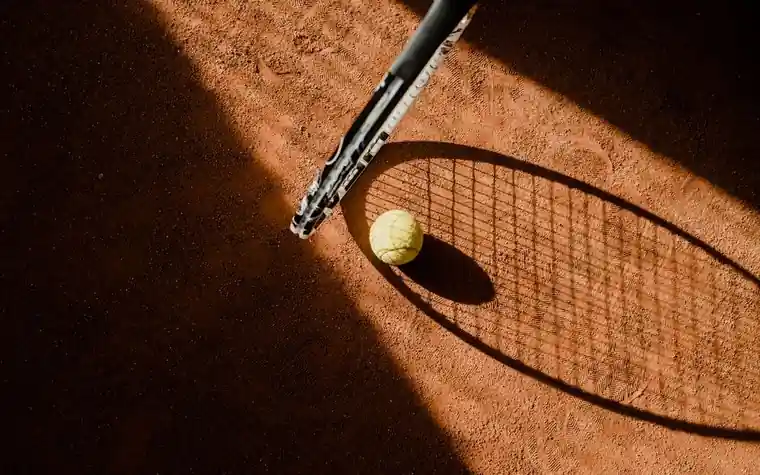 Tennis ball resting beside a racket on a sunlit clay court with the racket's shadow stretching across the surface