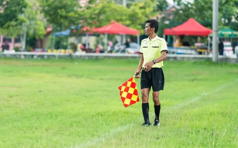 Soccer assistant referee in a yellow kit holding a checkered flag on a grass field