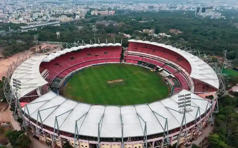aerial view of a large cricket stadium with a red and white curved roof surrounding a green oval field