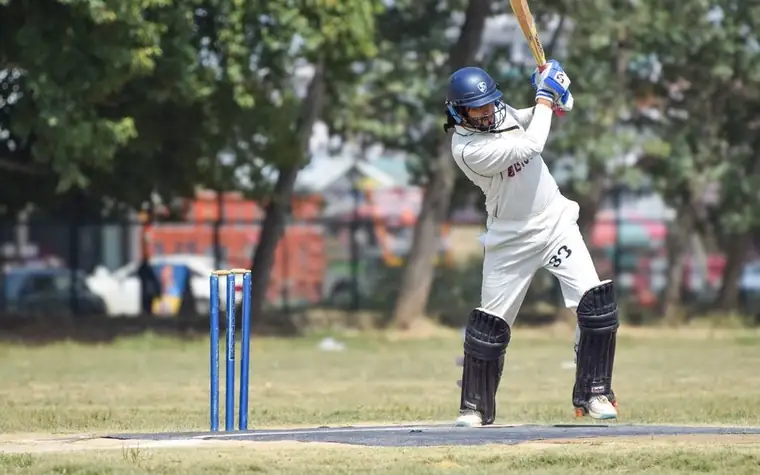 batsman in white playing a defensive stroke in front of blue stumps on a practice pitch with trees behind
