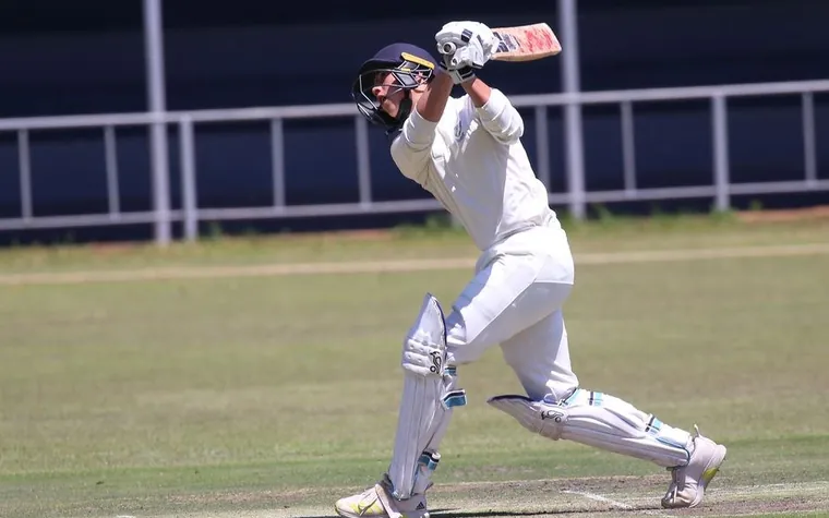 cricket batsman in white kit mid swing hitting a shot during a match with dark fencing and spectators behind