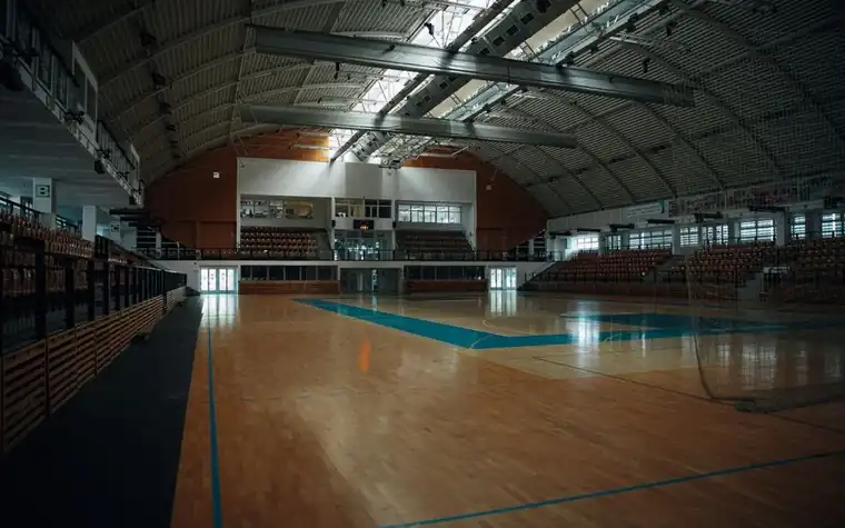 empty indoor basketball arena with polished hardwood floor, painted court lines and rows of bleachers under a steel roof