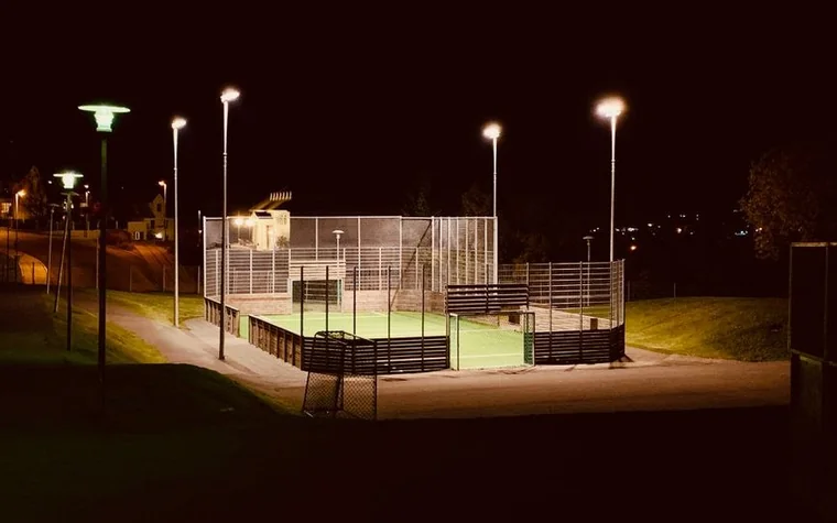 lit fenced football pitch with tall floodlight poles glowing against a dark night sky and a few bare trees
