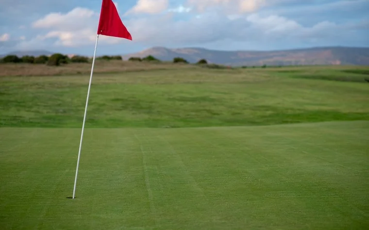 red flag on a pole in the middle of a mown green on a golf course with rolling hills and a cloudy sky behind