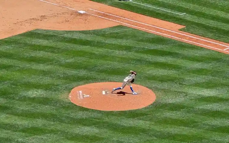 a pitcher in a white uniform at the end of his delivery on a dirt mound in the middle of a striped green baseball infield