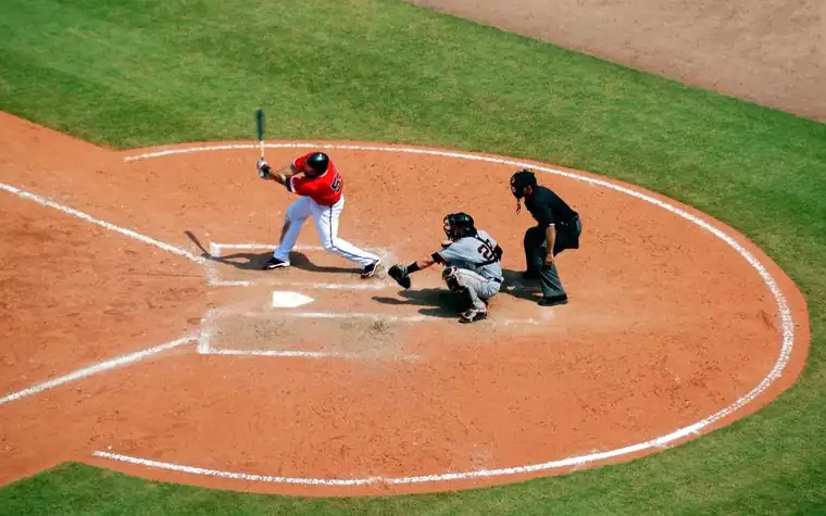 a batter in a red jersey swinging at home plate while a catcher and an umpire crouch behind him on a brown dirt infield