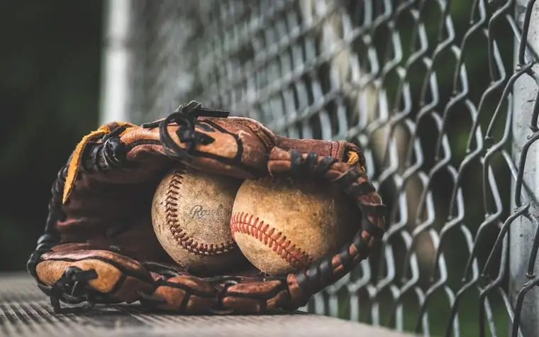 a worn brown leather baseball glove holding two aged baseballs on a bench in front of a chain-link fence