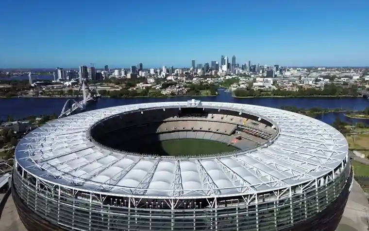 aerial view of a large oval stadium with a white roof beside a river and a city skyline under a clear blue sky