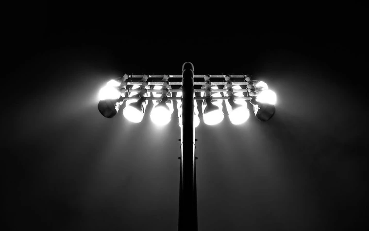 black and white close-up of a bank of bright stadium floodlights on a tall mast against a dark sky