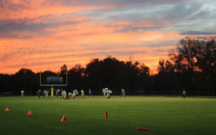 wide view of a football field under an orange and pink sunset sky with players gathered at one end and trees behind the stands