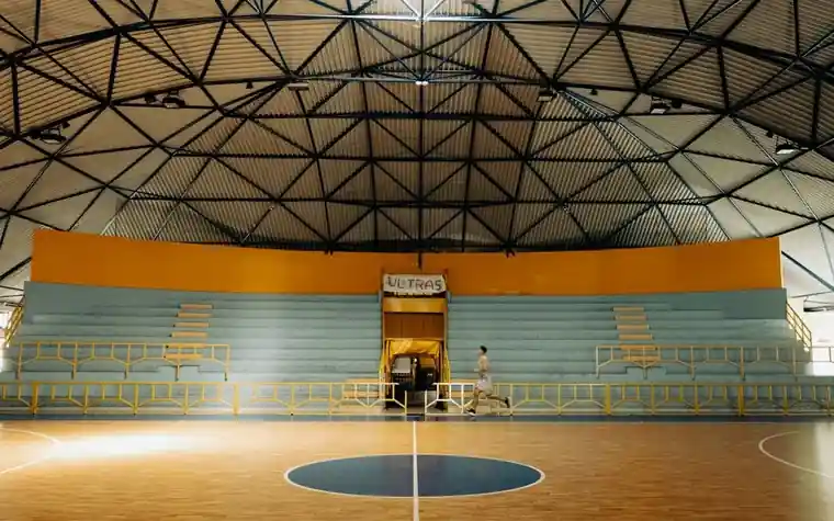empty indoor basketball arena with a steel truss roof, rows of bleachers and a lone person running across the polished court