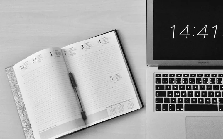 black and white photo of an open daily planner with a pen resting on its page beside the keyboard of a laptop showing the time