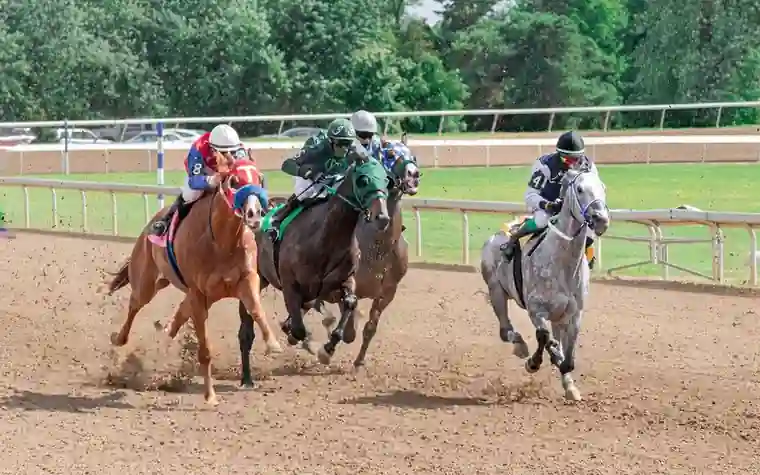 three jockeys in colored silks racing three horses side by side around a dirt track turn with a rail behind them