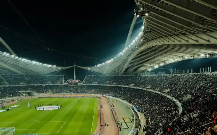 a floodlit stadium at night with packed curved stands around a green pitch seen from high in the upper tier