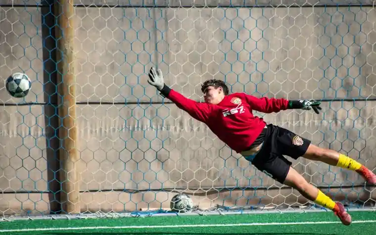 a goalkeeper in a red top and black shorts diving flat out through the air along a wall with a soccer ball on the ground behind him