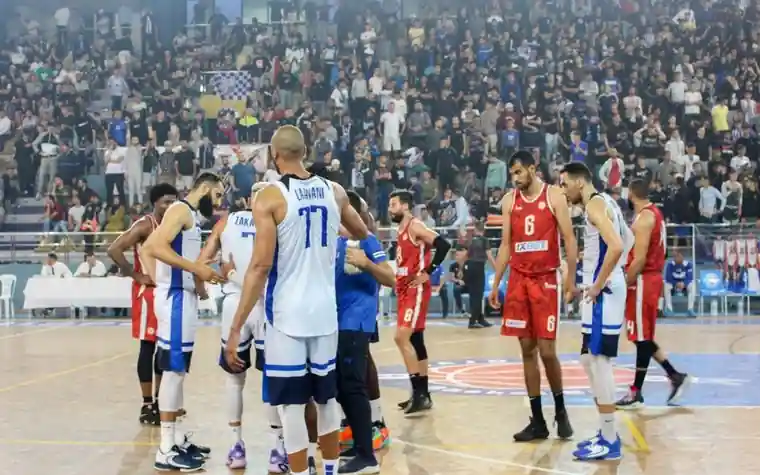 Basketball players from two teams in red and white jerseys huddle near the free-throw line as a packed stadium crowd watches from the stands