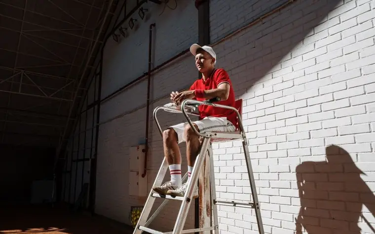 A tennis umpire in a red shirt and white cap sitting on a high umpire chair against a sunlit white brick wall