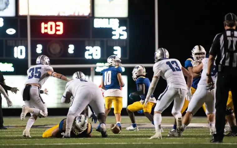 American football players and a referee on the field at night with a lit scoreboard showing the game clock and score in the background